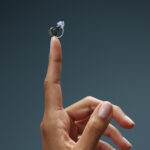 Tiny Oticon Zeal hearing aid held on fingertip.