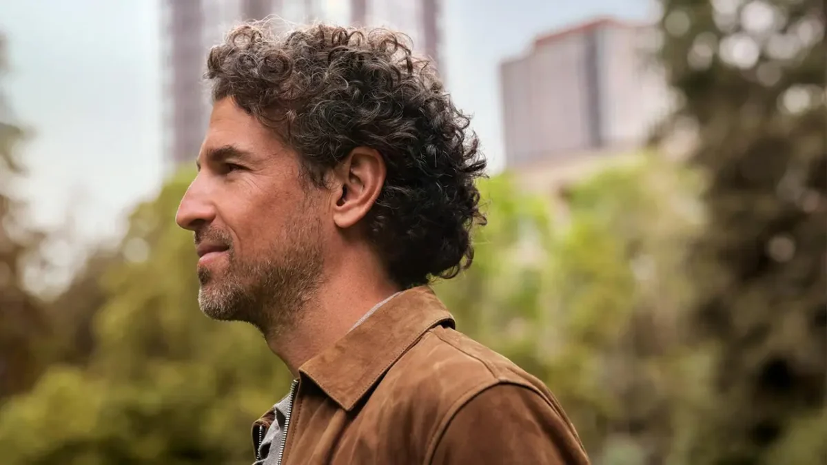 Side profile of a middle-aged man with curly dark hair wearing a discreet beige Oticon Zeal hearing aid in his right ear, outdoors with blurred greenery and buildings in the background.