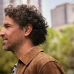 Side profile of a middle-aged man with curly dark hair wearing a discreet beige Oticon Zeal hearing aid in his right ear, outdoors with blurred greenery and buildings in the background.