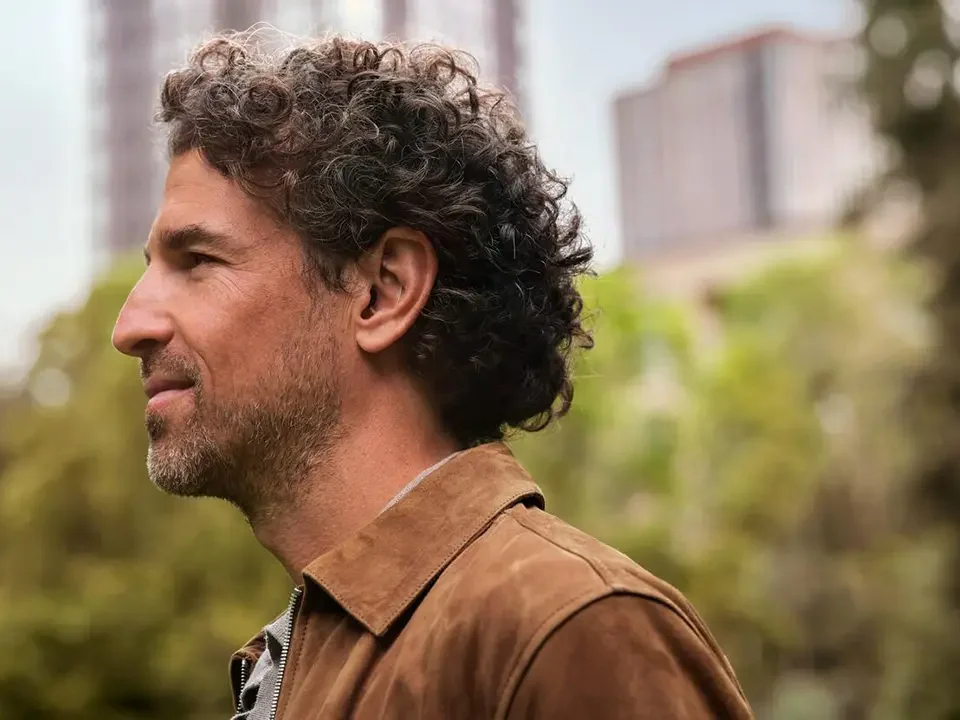 Side profile of a middle-aged man with curly dark hair wearing a discreet beige Oticon Zeal hearing aid in his right ear, outdoors with blurred greenery and buildings in the background.